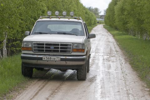 1993 Ford Bronco - A white 1990s Ford Bronco is parked on a muddy dirt road lined with trees. The vehicle has a roof rack with four auxiliary lights and a lic…