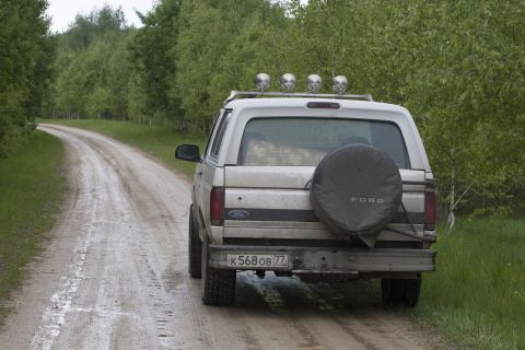 1993 Ford Bronco - The rear view of a silver Ford Bronco is shown parked on a wet, dirt road surrounded by trees. The vehicle has a spare tire mounted on the…