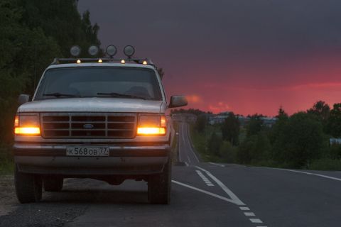 1993 Ford Bronco - A white Ford Bronco is parked on the side of a road at dusk, with its headlights on. The sky is a vibrant mix of dark purple and red.