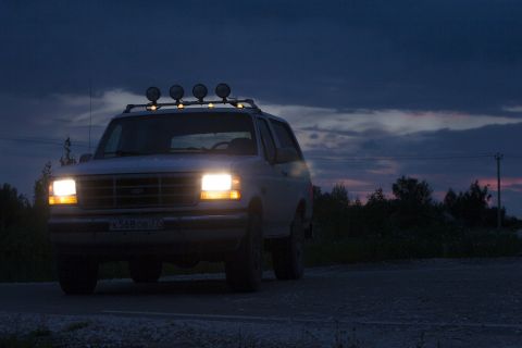 1993 Ford Bronco - A white Ford Bronco with its headlights on is parked on a gravel road at dusk. The vehicle has a roof rack with four auxiliary lights.