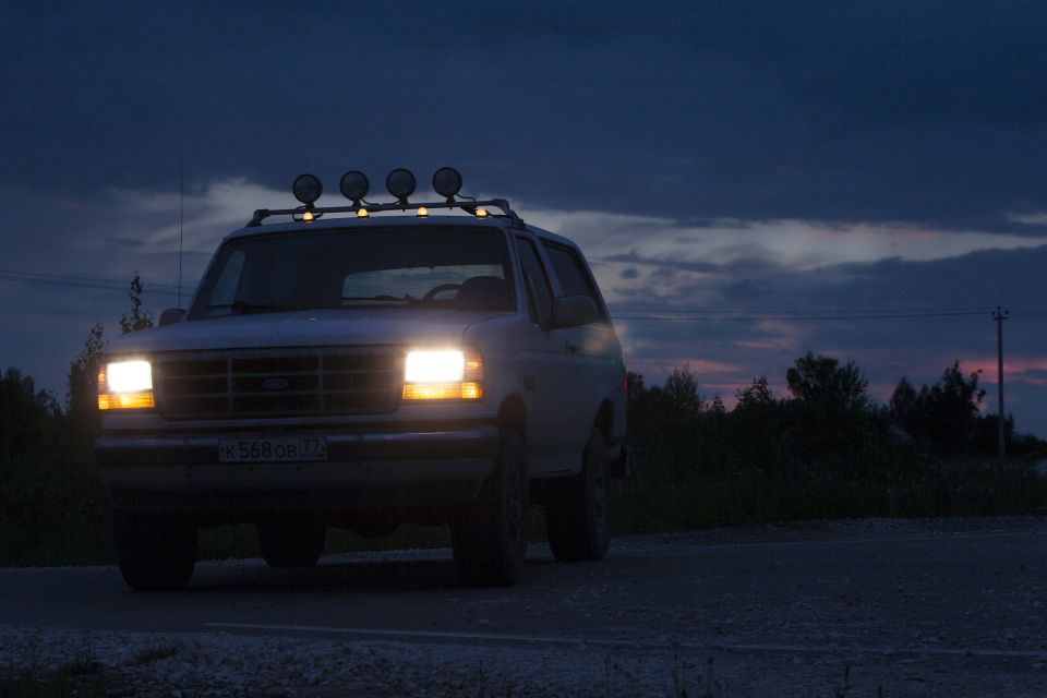 White 1993 Ford Bronco with headlights on and roof rack lights at dusk.