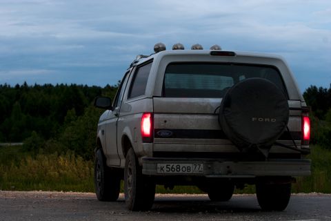 1993 Ford Bronco - The rear view of a white Ford Bronco is shown parked on a dirt road at dusk, with its taillights illuminated. A spare tire is mounted on th…