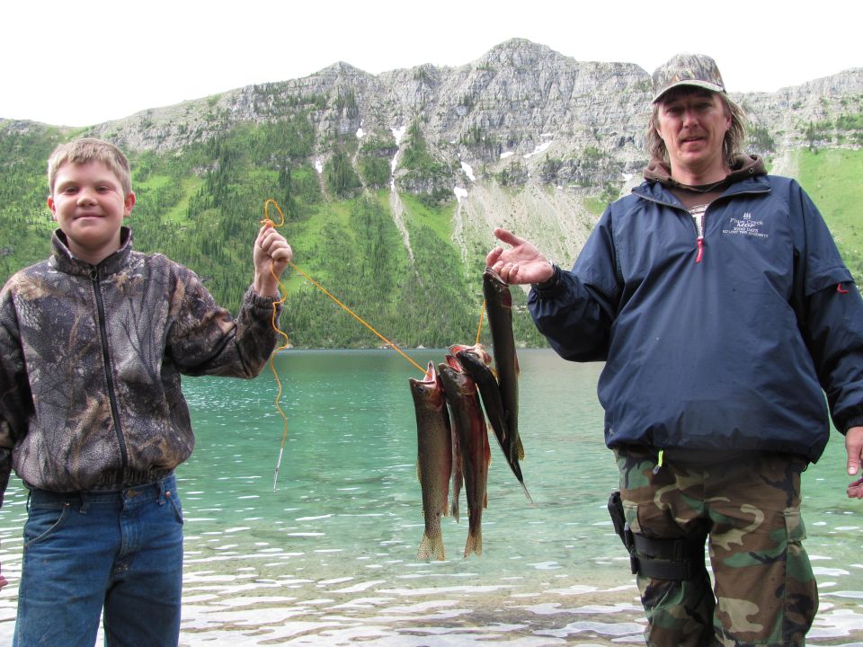 Two people holding up caught fish by a lake with mountains in the background.