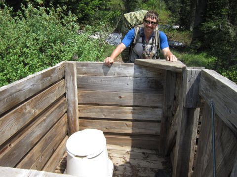 1974 Ford Ranchero - A man in sunglasses and a blue shirt stands behind a wooden outhouse with a toilet inside. He is wearing a backpack and leaning on the wood…