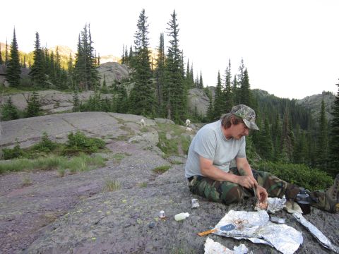 1974 Ford Ranchero - A man in camouflage pants and a baseball cap sits on a rocky outcrop, preparing food on aluminum foil. He is surrounded by pine trees and a…