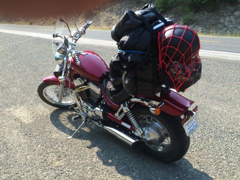 1974 Ford Ranchero - A dark red motorcycle is parked on the side of a road, loaded with luggage and a helmet secured with a red net. The motorcycle has a windsh…