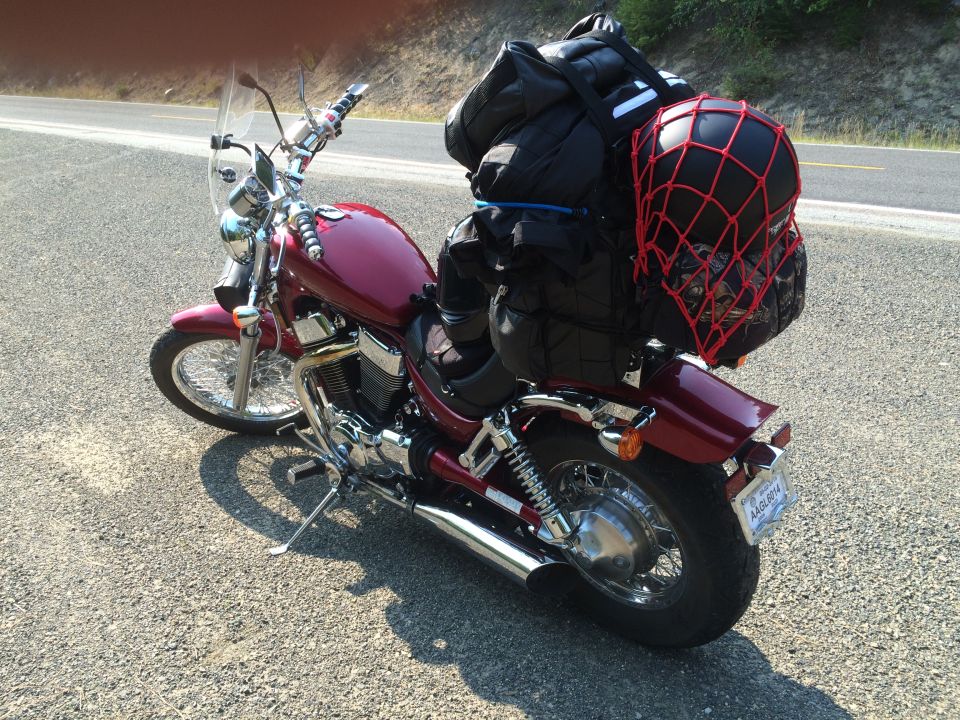 Dark red motorcycle loaded with luggage and a helmet, parked on a roadside.