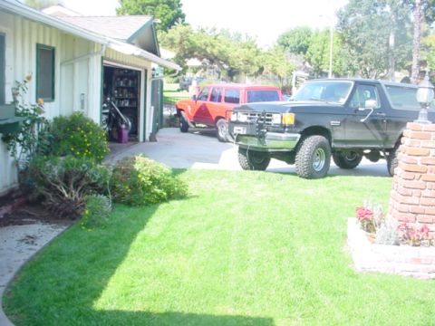 1988 Ford Bronco - Two SUVs are parked in a driveway next to a residential garage on a sunny day.