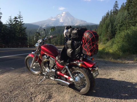 1974 Ford Ranchero - A red motorcycle is parked on a gravel shoulder next to a road, with a large mountain and forest in the background. The motorcycle is equip…