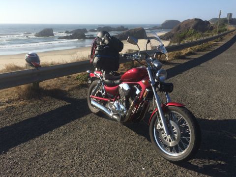 1974 Ford Ranchero - A red motorcycle is parked on the side of a road overlooking a beach with large rock formations in the ocean. A helmet rests on a guardrail…