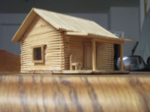 1974 Ford Ranchero - A close-up, low-angle shot of a miniature log cabin model made from toothpicks. The cabin has a porch with a chair and a small window.