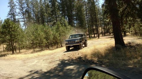 1995 Ford Bronco - A dark 1992 Ford Bronco kicks up dust as it drives along a dirt road through a wooded area.