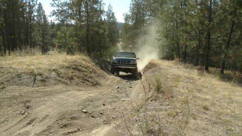 1995 Ford Bronco - A Ford Bronco kicks up dust as it navigates a narrow dirt path surrounded by trees.