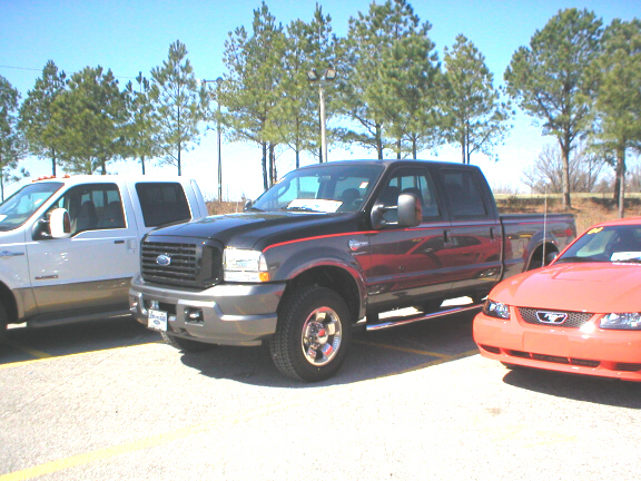 Dark red and gray Ford Super Duty crew cab pickup parked between a white truck and orange Mustang