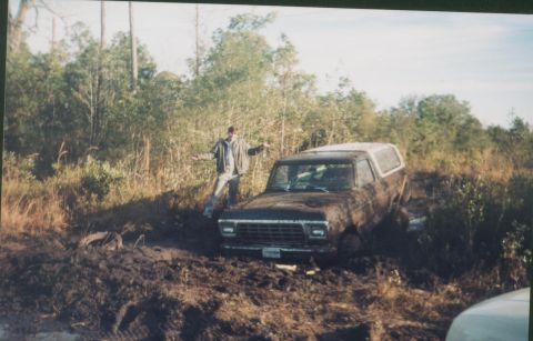 1993 Ford Bronco - A person stands beside a mud-caked SUV that appears to be stuck in a swampy, wooded area.