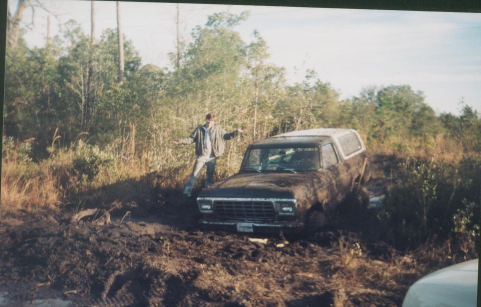 Mud-covered Ford Bronco stuck in deep mud with a person standing beside it