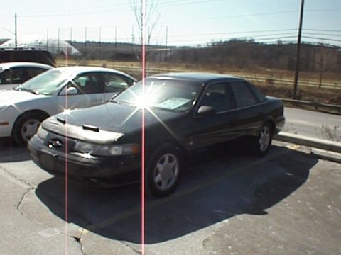 1999 Ford Taurus - A dark 1999 Ford Taurus sedan is parked in a lot, partially covered by a car bra. Another white car is visible to the left.