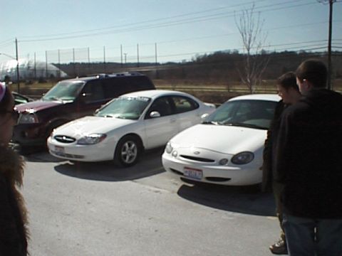 1999 Ford Taurus - Two white Ford sedans and a dark SUV are parked in a lot under a clear sky.