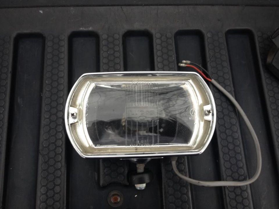Close-up of a rectangular headlight with a chrome bezel and wires attached, resting on a textured surface