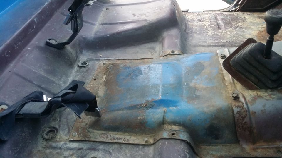 Truck interior floor with rust and blue paint, showing seatbelt anchors and a shifter boot