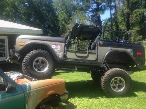 1972 Ford Bronco - lifted early Ford Bronco with two-tone tan and black paint and oversized tires sits on a lawn near a rusty fender.