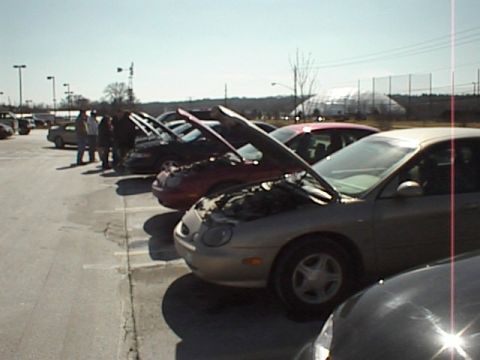 1999 Ford Taurus - A line of cars, including a silver 1999 Ford Taurus with its hood open, are parked in a lot under a bright sky.