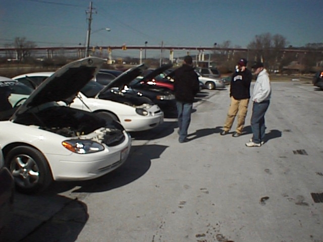 Several Ford Taurus cars parked with hoods open, three people standing nearby