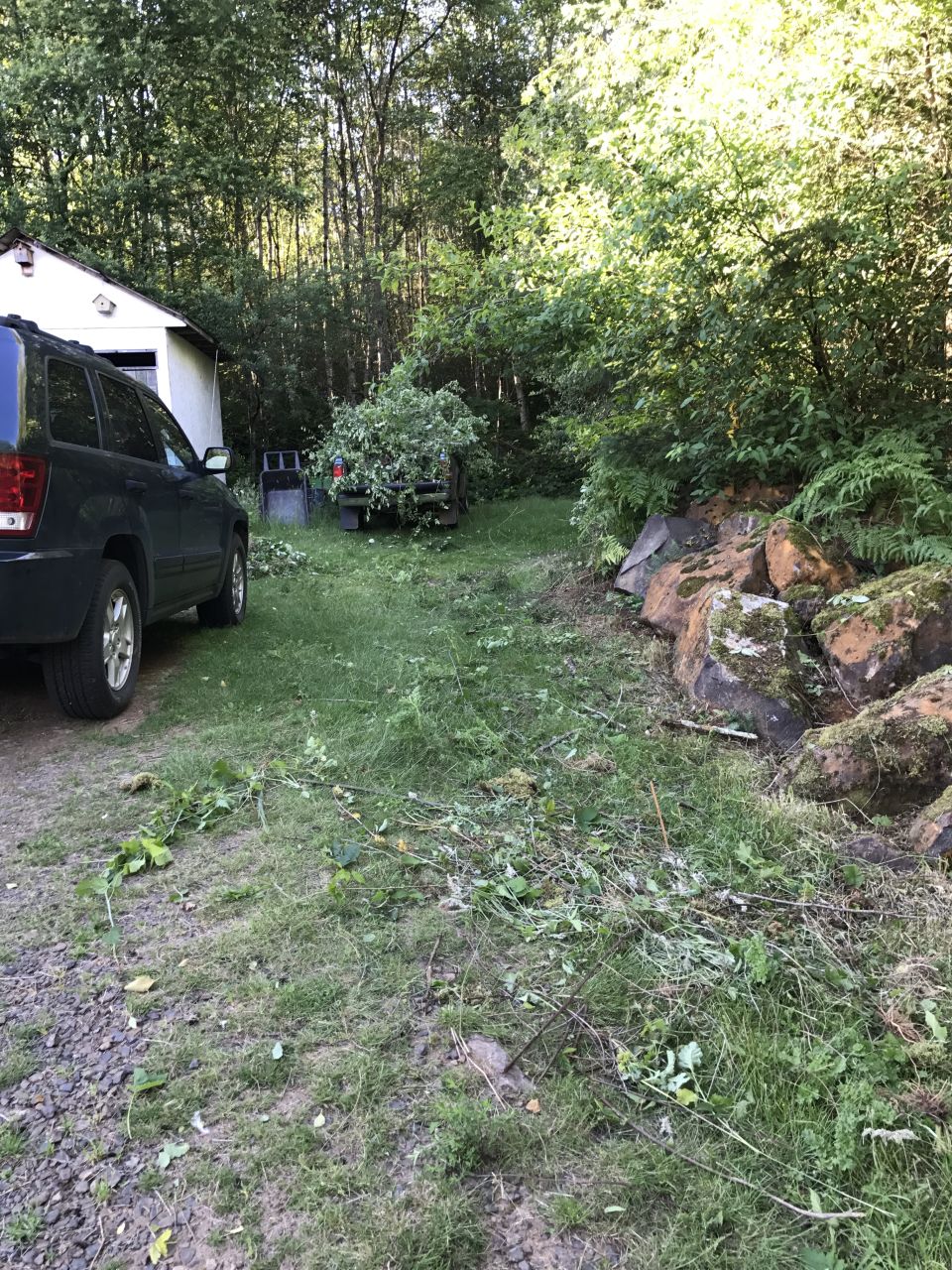 Dark SUV parked next to a garage, with a trailer full of branches behind it. Rocks and trees in the background