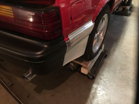 1983 Ford Mustang - Close-up of a red car's rear end, showing the bumper and quarter panel area, which is undergoing bodywork.