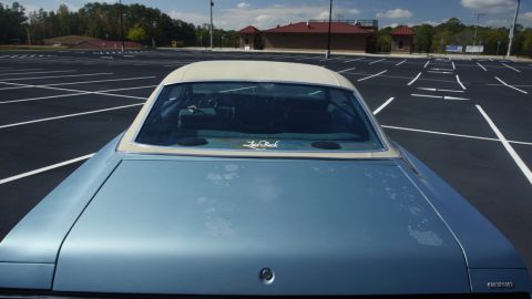 1975 Chevrolet Chevelle - Rear view of a light blue convertible with a white top, parked in an empty lot under a partly cloudy sky.