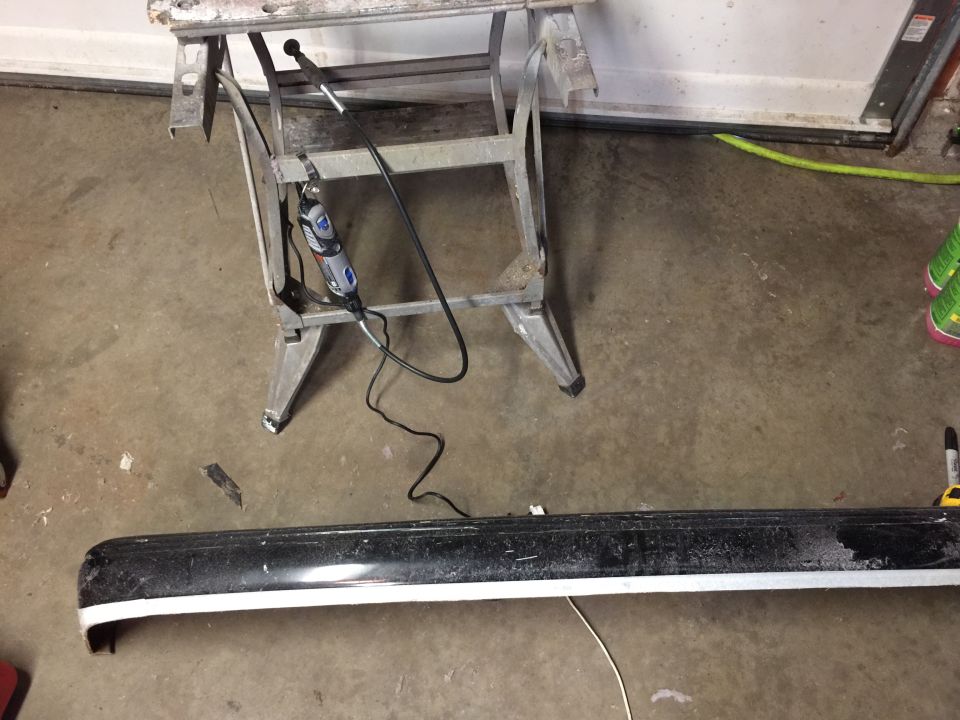 Black car bumper with a white stripe, resting on a concrete floor near a sawhorse holding a Dremel tool