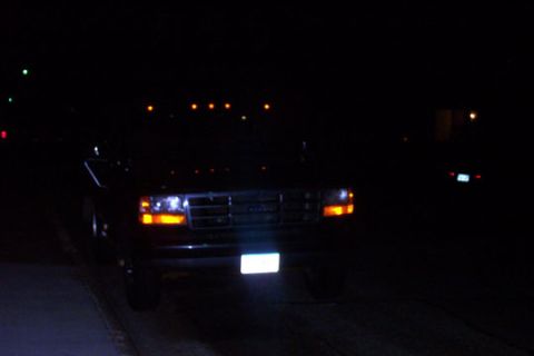 1995 Ford Bronco - A dark Ford F-Series truck is seen from the front at night, with its headlights and license plate illuminated.
