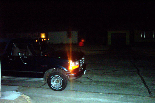 Dark 1990s Ford SUV at night on concrete lot, amber turn signal lit, white-letter tires