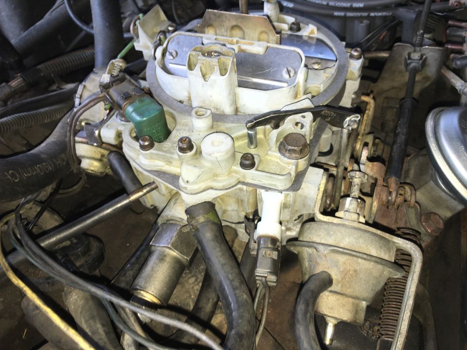 Close-up of a vehicle carburetor with hoses, wires, and linkages.