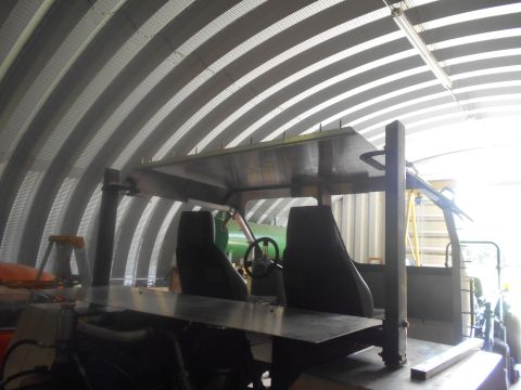 1992 Ford Bronco - Interior view of a custom-built vehicle with two black seats, a steering wheel, and a metal roof structure, inside a Quonset hut with corru…