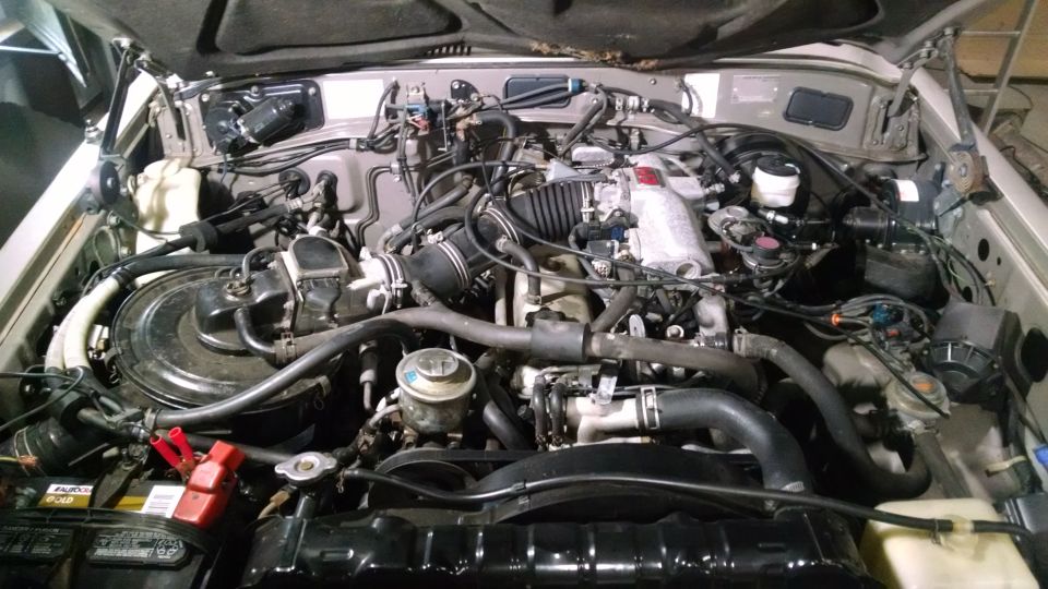 Open engine bay of a silver SUV showing inline engine, hoses, and battery with red jumper clamps