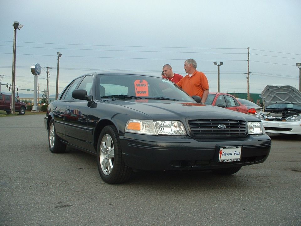 Dark Ford Crown Victoria with WAS/NOW window sticker on a dealer lot, two men beside it