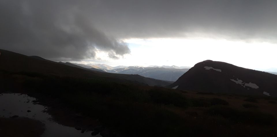 Mountain landscape with dark storm clouds overhead and a bright sky in the distance. There are puddles on the ground in the foreground