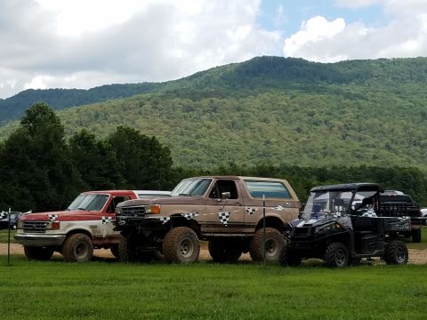 1988 Ford Bronco - Three off-road vehicles, including a red and white Ford pickup and a brown Ford Bronco, are parked on a grassy field with a wooded mountain…