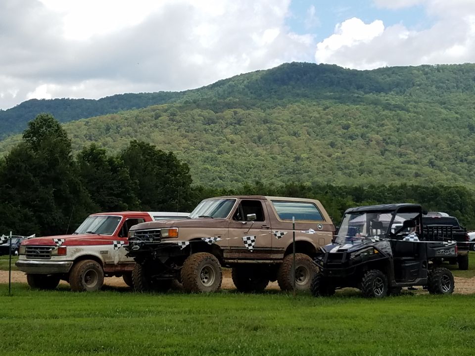 Red and white Ford pickup, brown Ford Bronco, and black UTV parked on grass with mountains behind.