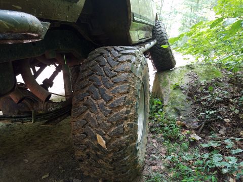1988 Ford Bronco - A close-up, low-angle shot of the muddy tires and undercarriage of a 4x4 vehicle navigating a rocky, wooded trail. The vehicle's large, agg…
