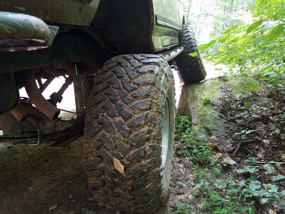 Muddy off-road tires of a 4x4 vehicle climbing over rocks on a wooded trail.