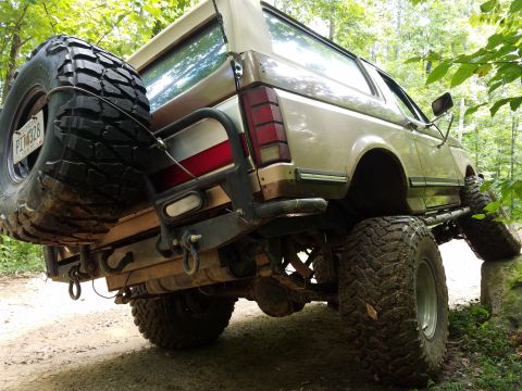 1988 Ford Bronco - The rear driver's side of a lifted, tan 1980s Ford Bronco is shown at a low angle, with its spare tire mounted on the back and large off-ro…