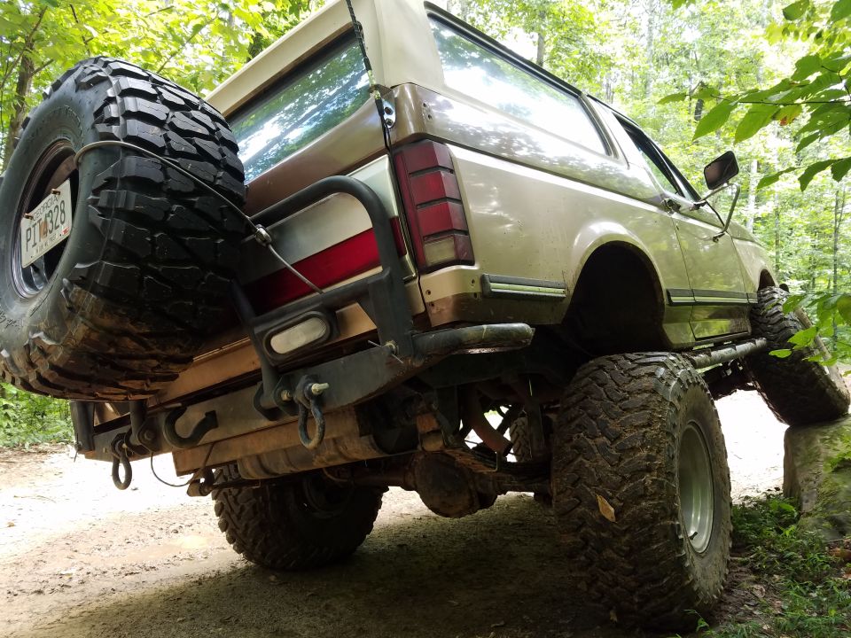 Lifted 1980s Ford Bronco, rear 3/4 view, with spare tire and large off-road tires on a dirt path.