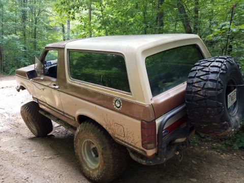 1988 Ford Bronco - A two-tone brown and tan Ford Bronco is parked on a dirt path in a wooded area. The SUV has large off-road tires and a spare tire mounted o…