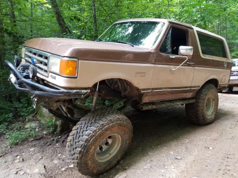 1988 Ford Bronco - A lifted, two-tone brown 1980s Ford Bronco with large off-road tires and a custom front bumper is parked on a dirt path in a wooded area. T…