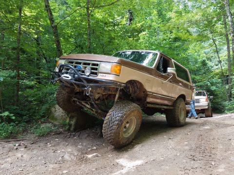 1988 Ford Bronco - A brown SUV with prominent tires navigates a dirt path surrounded by trees.