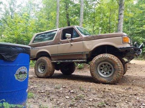 1988 Ford Bronco - This lifted Ford Bronco is parked on a dirt path surrounded by trees, with a blue barrel to its left.