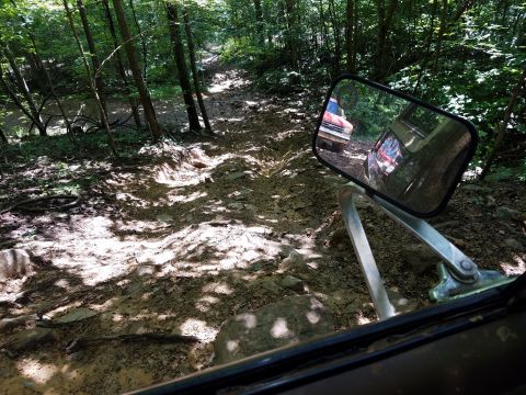 1988 Ford Bronco - A red truck navigates a challenging muddy trail, seen through the side mirror of another vehicle.