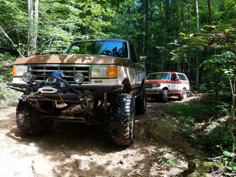 1988 Ford Bronco - Two lifted SUVs navigate a wooded trail, the lead vehicle a prominent winch and off-road modifications.